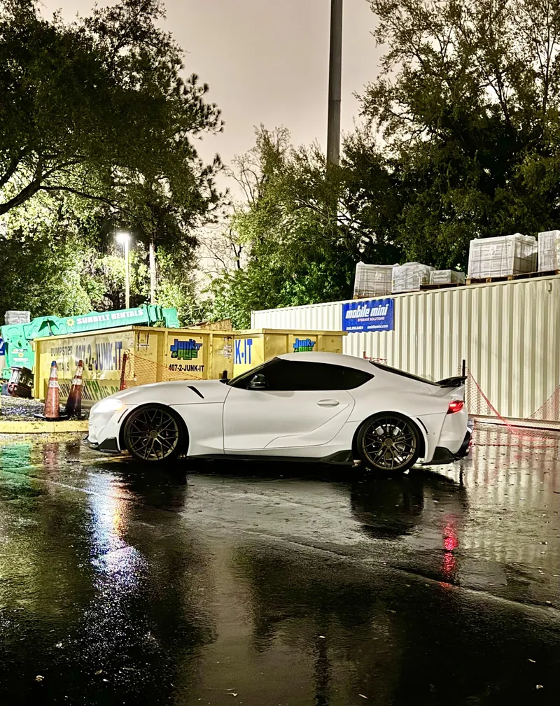 A white GR Supra on wet asphalt at night, lights reflected in the puddles