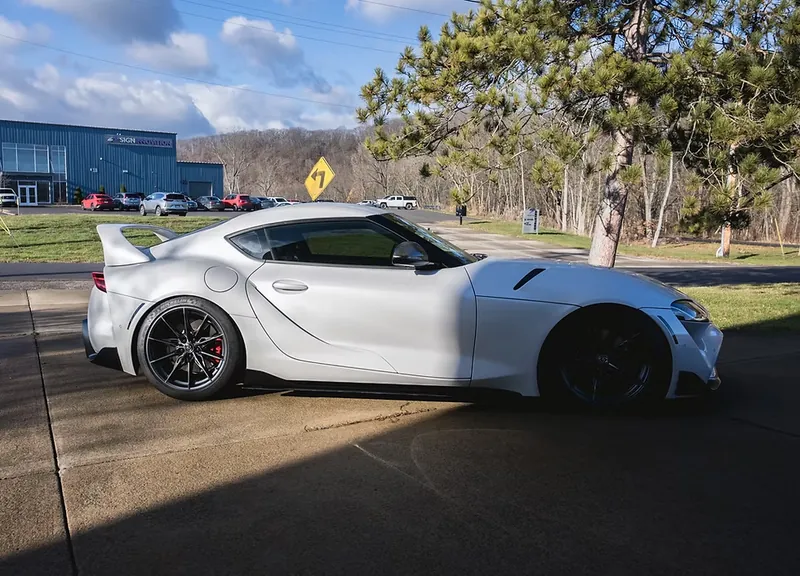 Side view of a white GR Supra with a rear wing, pine trees and blue sky behind